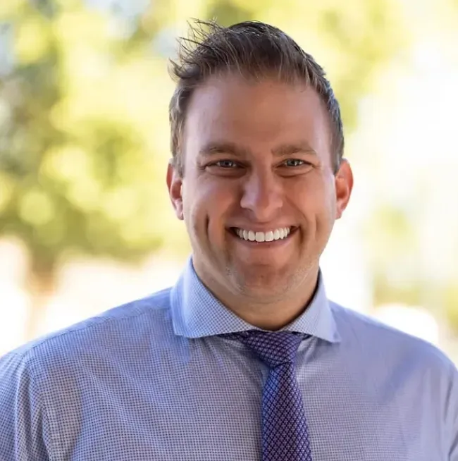 Smiling man with short hair wearing a light blue patterned shirt and tie.